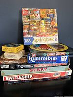 Photo of stacked vintage board games including Battleship, Sorry!, Rummikub, Battlestar Galactica, Chinese Checkers, and a completed 1000-piece Springbok puzzle on top.