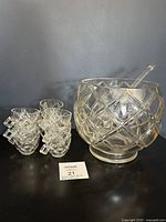 Glass punch bowl with diamond block pattern, ladle inside bowl, nine matching cups grouped beside it on black surface.