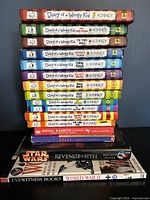 Stack of 16 children's books featuring 12 Diary of a Wimpy Kid titles along with additional books on various subjects. Vertical view showing colorful spines with author Kinney prominent on Diary of a Wimpy Kid titles and additional titles below in the pile.