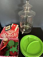 Photo showing the glass drink dispenser with metal stand and spigot, green vintage cookie cutters in red gingham box, and Pimpernel coasters on top of a red and white placemat.