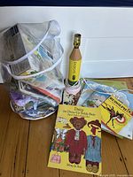 Photo of a large clear mesh bag filled with assorted arts and crafts supplies, several vintage children's books and records are placed next to it on a floor.