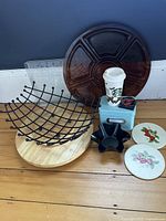 Kitchenware lot consisting of wooden lazy Susan, wire fruit basket, serving platter, recipe tin with Starbucks cup, tortilla bakers, trivets, magnet, and decorative trays arranged on wooden floor.
