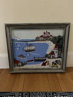 Framed vintage crewel embroidery featuring a coastal scene with sailboat, houses, lighthouse, dock, and birds on blue water.