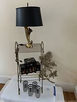 Full lot arrangement showing lamp on top of metal side table, Japanese musical jewelry box on table shelf, four bar glasses on bottom shelf, and floral wall plaque leaning behind