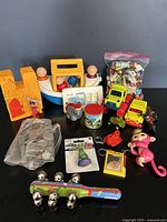 Lot of small toys including Tupperware Noah's Ark boat with wooden figures, multiple small toy figures in ziplock bag, two yellow toy trucks, pink Fingerling monkey, small elephant, toy castle, and multicolored toy with silver balls on front of gray background