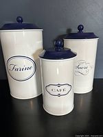 Front view of three white metal canisters with blue lids, labeled in French 'Farine', 'Sucre', and 'Café'