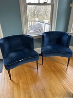 Front view of two blue barrel-back chairs with tapered dark wooden legs, placed on wood floor in front of window.