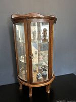 Photo showing the small vintage wooden curio cabinet closed, displaying curved glass panels, four legs, and visible knick knacks inside on glass shelves with a mirrored back.