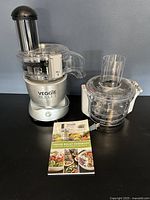 Full view of the assembled Veggie Bullet machine with motor base, primary food processor attachment, cylindrical feeding tube with black pusher, and the Veggie Bullet cookbook placed in front on a table.