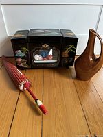 Front view of lot showing large black lacquer Japanese music box with colorful painted scenes and display window, red Asian parasol folded beside it, and brown midcentury ceramic pitcher.
