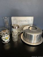 Photo showing overall lot including aluminum serving tray, cake holder, decorative tin canister, glass canning jar, glass pitcher, and liqueur bottle arranged together.