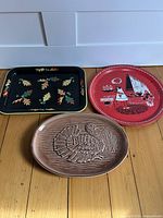 Three vintage trays arranged on a wooden floor: black metal autumn leaf tray, red round BBQ tray, and brown ceramic turkey platter.