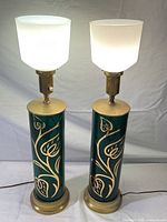 Pair of emerald green glass cylindrical table lamps with gold vine and leaf painted decoration, white ribbed glass shades attached, shown lit and unlit.