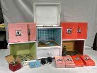 Front view of several American Girl Minis light boxes arranged with some boxes stacked. Two rooms are visible with wallpaper and miniature furniture inside. Boxes in red and pink colors labeled A*G Minis placed around.