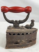 Side view of the antique coal iron showing rusted cast iron body, red wooden handle and rooster-shaped latch