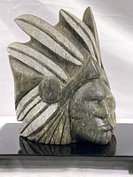 Close-up side view of heavy stone carving of Mayan warrior head showing feathered headdress and facial features. Mounted on black painted wood stand.
