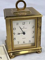 Front view of Hamilton quartz clock showing white face, black numerals from 1 to 12, and black hour, minute, and second hands. Clock in brass-tone casing with top circular handle.