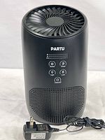 Front view of the black Partu BS-03 air purifier showing the control panel with buttons and indicators, and attached power cord and plug.