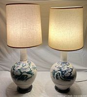 Pair of vintage table lamps illuminated showing ivory beige textured shades and hand-painted porcelain bases with Asian dragon and floral motifs.