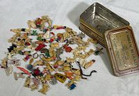 Overview of various small vintage plastic charms spread out next to an open vintage metal Cadillac Automobile Bulb Kit tin box on a white cloth background, showing the breadth of the collection and storage container.