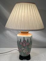Full view of the vintage table lamp illuminated, showing ceramic base with hand-painted iris flowers and pleated cream lampshade.