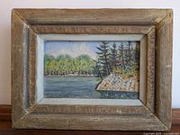 Frontal view of the entire framed landscape painting showing the river, trees and rocks.