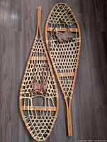 Full view of a pair of wooden antique snowshoes with traditional lacing and leather bindings, displayed on wooden floor.