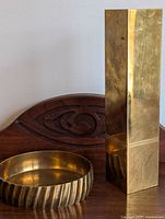 Tall rectangular brass vase and round brass bowl placed on wooden surface against wall, showing wear and tarnish.