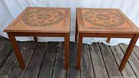 Front view of two square wooden side tables with decorative tile tops and four legs, sitting on wooden floor and white backdrop.