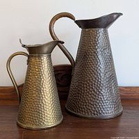 Two hammered metal jugs, one larger copper with dark patina, one smaller brass with golden tone, placed on wooden surface.