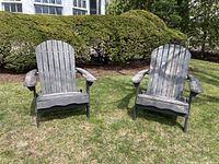 Two wooden Adirondack chairs placed outdoors on lawn with green bushes in background. Chairs have worn paint finish.