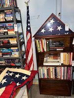 Image showing three American flags: one folded inside a wooden triangular case on a bookshelf, one loose folded flag on a chair, and one flag mounted on a pole with an eagle topper.