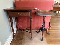 Photo showing two antique wooden tables placed side by side - a rectangular end table with drawer and a round pedestal side table with metal gallery rim around the top.