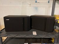 Front view of two black Bose speakers side by side on a gray shelf.