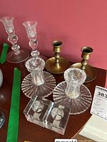 Photo showing 2 brass candle holders, 2 ribbed glass candle holders with broad flat base, and 2 small clear square block candle holders on wooden surface