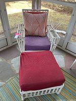 Photo showing white painted rattan chair with purple seat cushion and red and white geometric patterned throw pillow alongside matching painted rattan footstool with red cushion.
