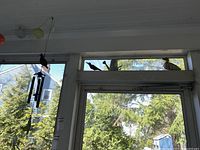 View of two metal wind chimes hanging on porch ceiling, with colored paper/fabric orb string lights and decorative bird sculptures on the window ledge above door, showing porch and outside trees.