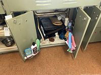 Photo of cabinet showing cooling racks, cookie sheets, wooden cutting board, various cleaning bottles, and sponges