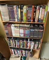 Four shelves of DVD cases in a wooden shelving unit, assorted titles and sets.