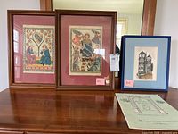 Three framed prints on a wooden table: two medieval-themed prints with a crowned figure and attendants, and one architectural drawing of a tower.