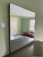 Front view of a square framed mirror resting against a pale green wall, reflecting parts of the room including a window and several furniture pieces.