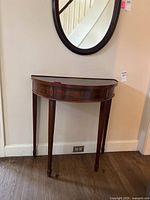Front side view of the semi-circular wooden hall table placed against a cream-colored wall under a round mirror. Shows drawer front and tapered legs.