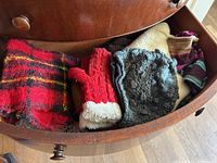 Drawer containing assorted mittens, hats, and scarves including a red and black plaid scarf, red mitten with white trim, grey mitten, beige and striped hats.