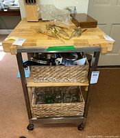 Front view of metal kitchen cart with butcher block top and two woven baskets in lower shelves, some kitchen utensils on the top surface including cutlery and serving spoons, knife block in background.