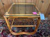 Front view of the square rattan coffee table showing the glass top and rattan lower shelf with a carpeted floor in the background.