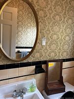 Oval gold framed wall mirror mounted above sink with tiled wall and vintage wallpaper background. Shows some wear on frame finish.