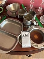 Photo showing multiple pewter and metal items including a divided serving dish, creamer, small cups, beer mug, and wine bottle coaster on a wooden table.