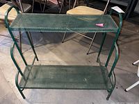 Front view of green metal two-tier plant stand showing two mesh shelves and curved side supports.