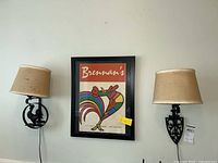 Two metal electric wall sconces with rooster designs and beige fabric shades flanking a framed Brennan's poster on a wall.