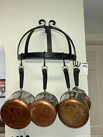 Front view of four copper bottom pots hanging on black metal pot rack mounted on wall, showing copper patina underside and black handles.